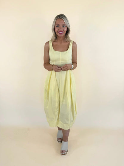 Woman wearing butter yellow midi dress with tulip hem, square neckline, and flowy silhouette styled with neutral slide sandals