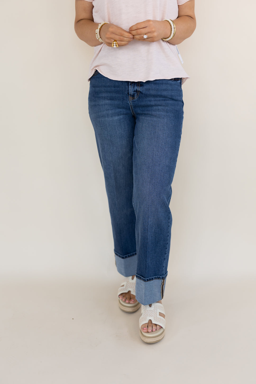 A person wearing blue straight-leg jeans with rolled cuffs, paired with a light pink t-shirt and white slide sandals, standing against a plain off-white studio background.