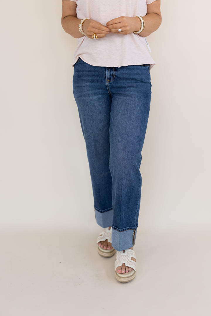A person wearing blue straight-leg jeans with rolled cuffs, paired with a light pink t-shirt and white slide sandals, standing against a plain off-white studio background.