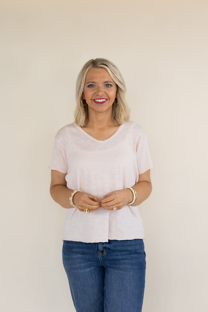 A smiling blonde woman wearing a light pink short-sleeve V-neck top and blue jeans, standing against a plain beige background.