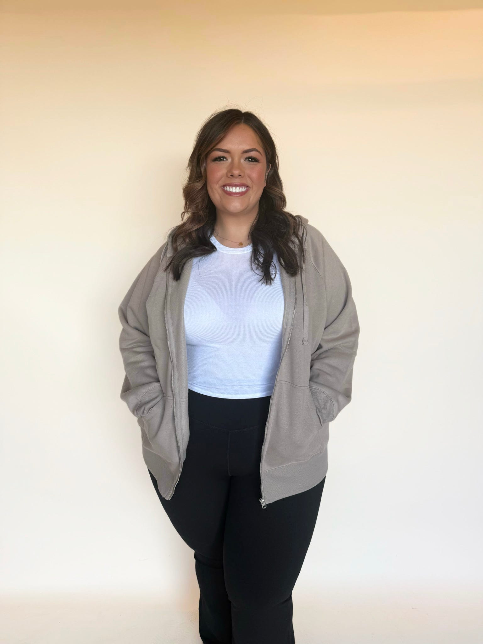 Woman modeling a taupe zip-up hoodie over a white top, hands in pockets, smiling in soft natural lighting.