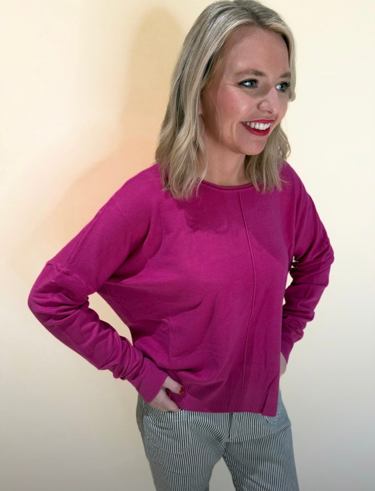 Side angle of a woman in a bright fuchsia sweater and striped pants, smiling and looking off camera with hands on hips, set against a light neutral backdrop.