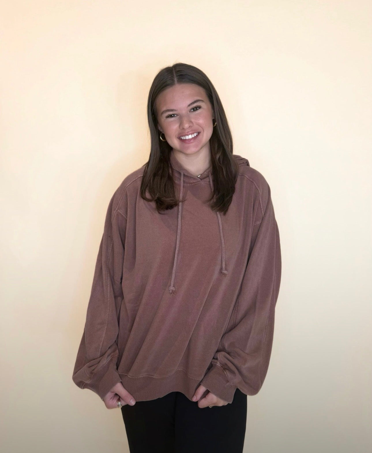 Woman wearing a loose mocha brown hoodie with drawstrings and black pants, smiling with hands lightly tucked at the hem, standing in front of a cream background.