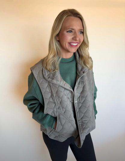 Woman wearing a gray quilted vest over a green sweater against a plain background