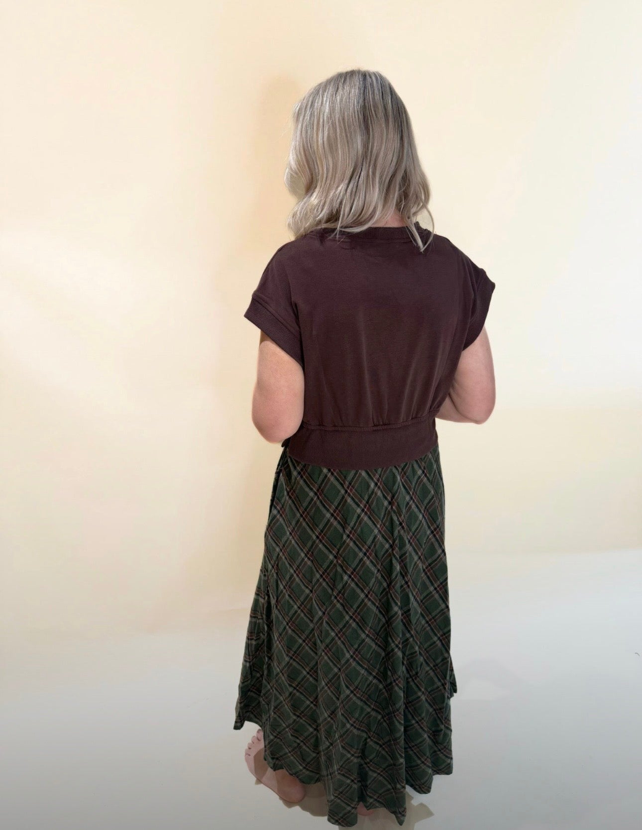 Back view of brown short-sleeve top with fitted waistband paired with flowy green plaid maxi skirt, model standing barefoot against a neutral background.