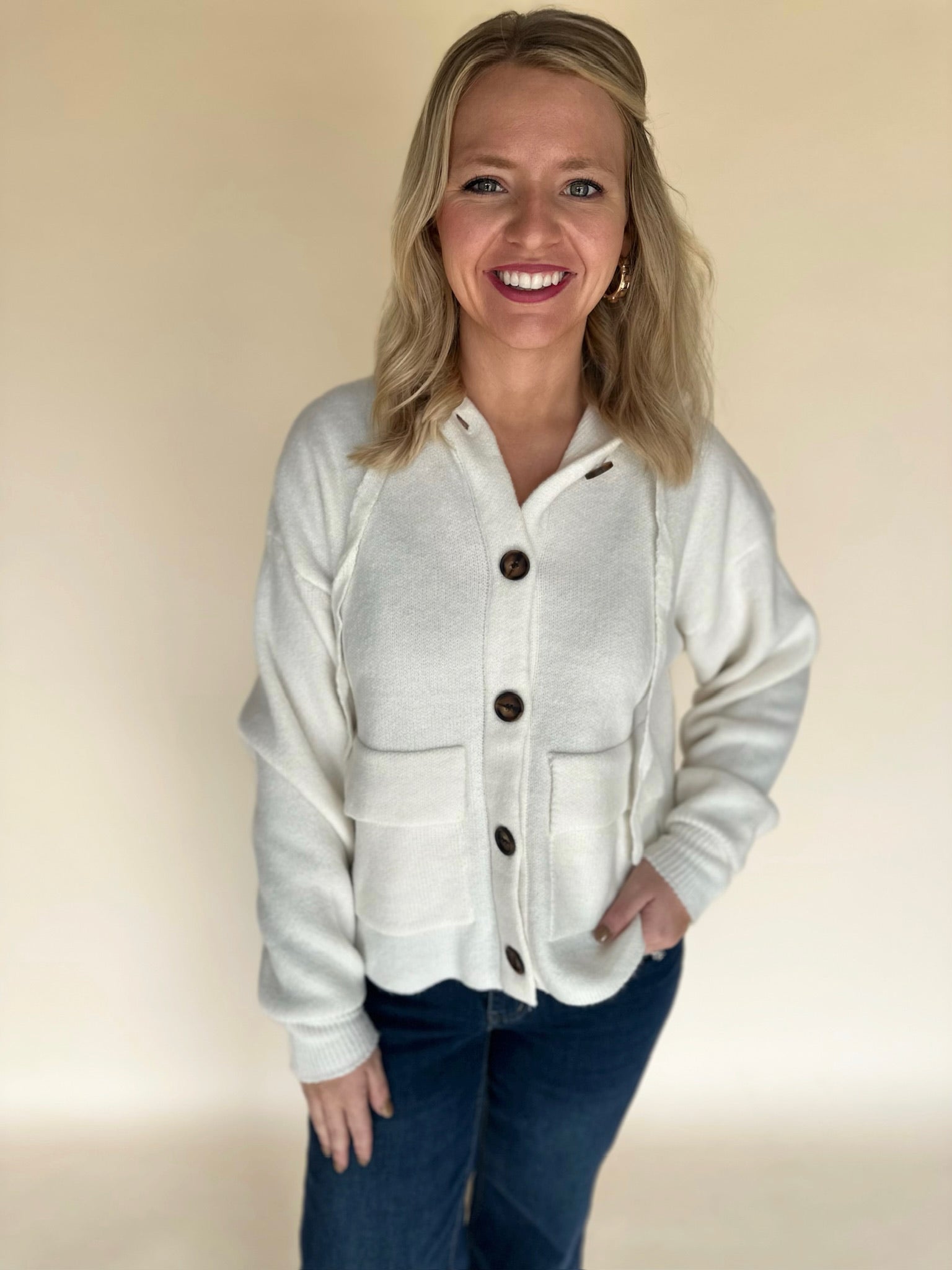 Woman wearing a cream knit button-front hoodie cardigan with two front pockets, styled with dark denim jeans, smiling and facing the camera against a neutral background.