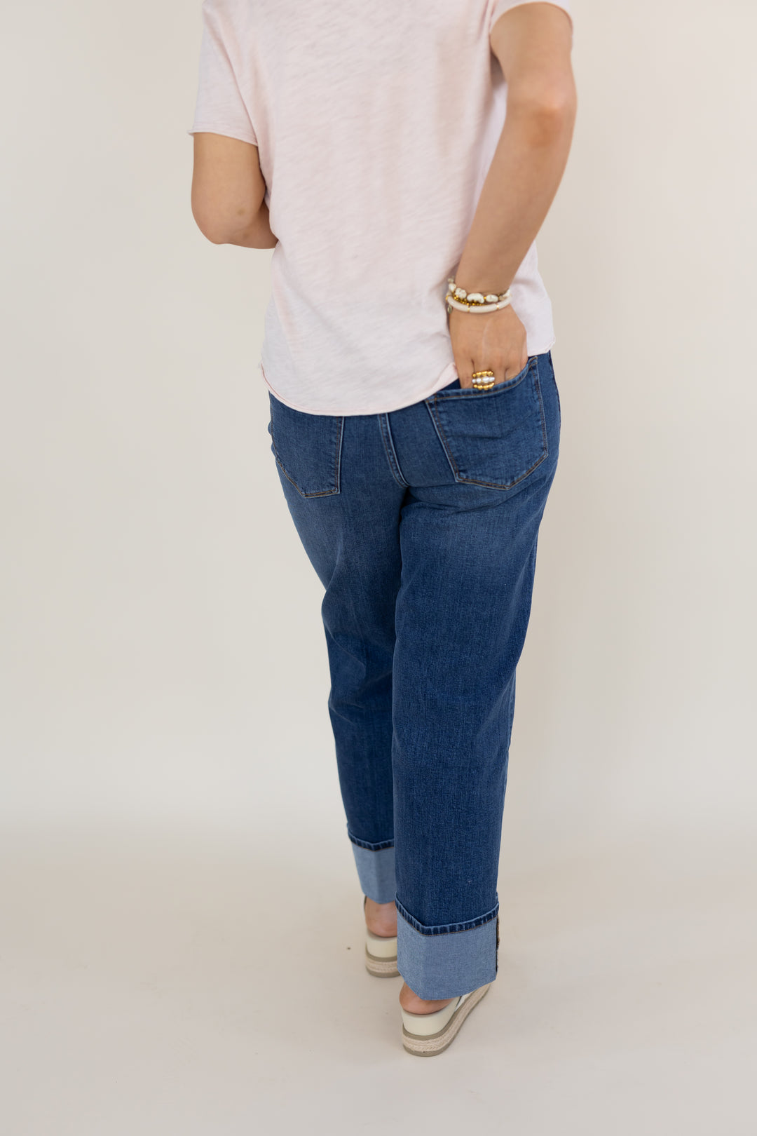 A person shown from the back wearing blue cuffed jeans, a light pink t-shirt, and beige-and-white platform sandals against a plain light gray studio background.