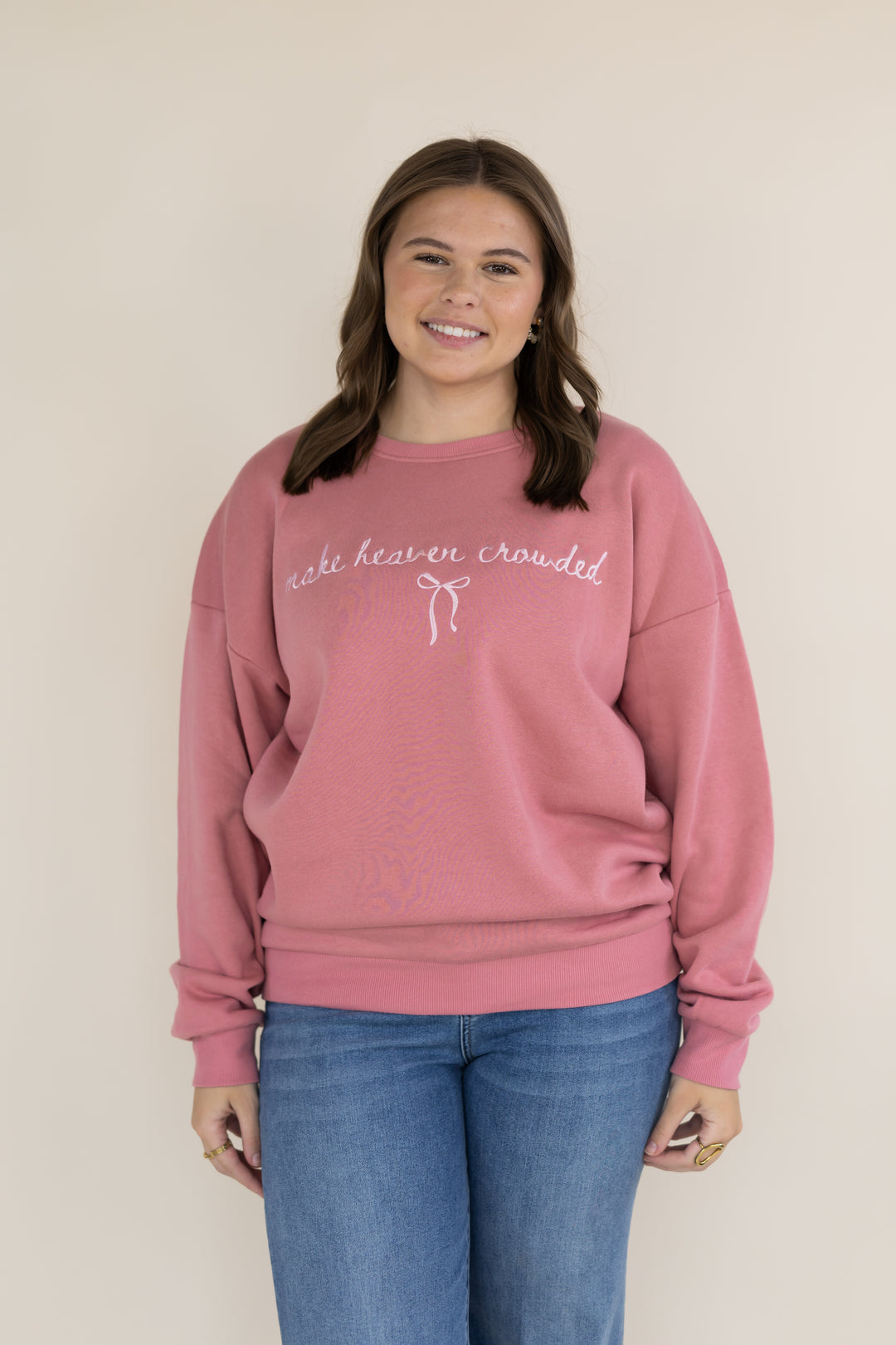 A woman wearing a dusty rose pink crewneck sweatshirt with white cursive embroidery that reads &