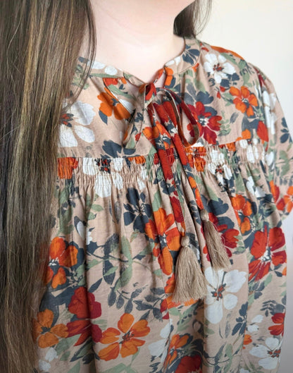 Close-up of floral smocked babydoll top showing tie-neck and beige tassel details in rust, navy, white, and olive floral print.