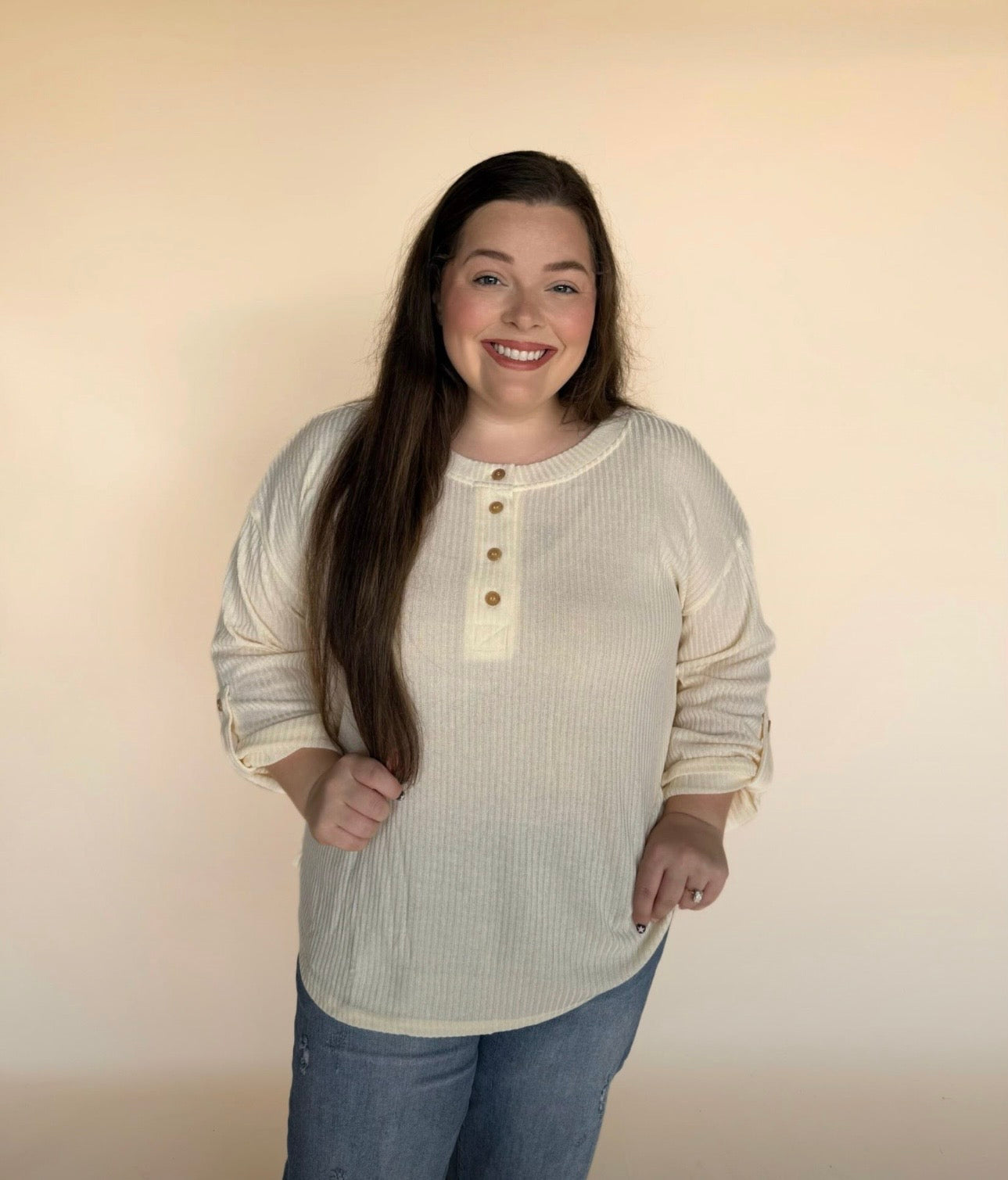 “Woman modeling a cream ribbed long-sleeve Henley with button neckline, standing against a light neutral background and smiling.