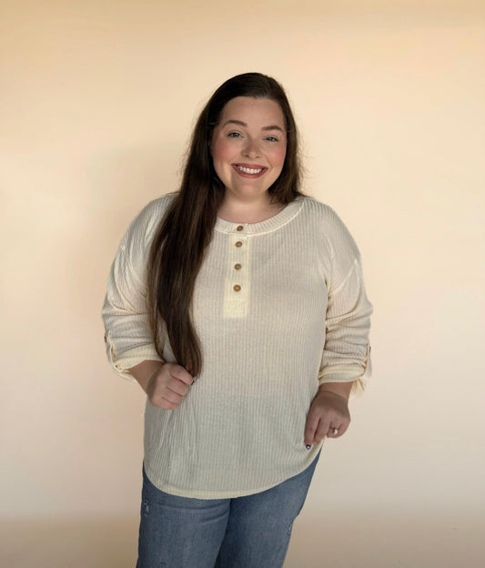 “Woman modeling a cream ribbed long-sleeve Henley with button neckline, standing against a light neutral background and smiling.
