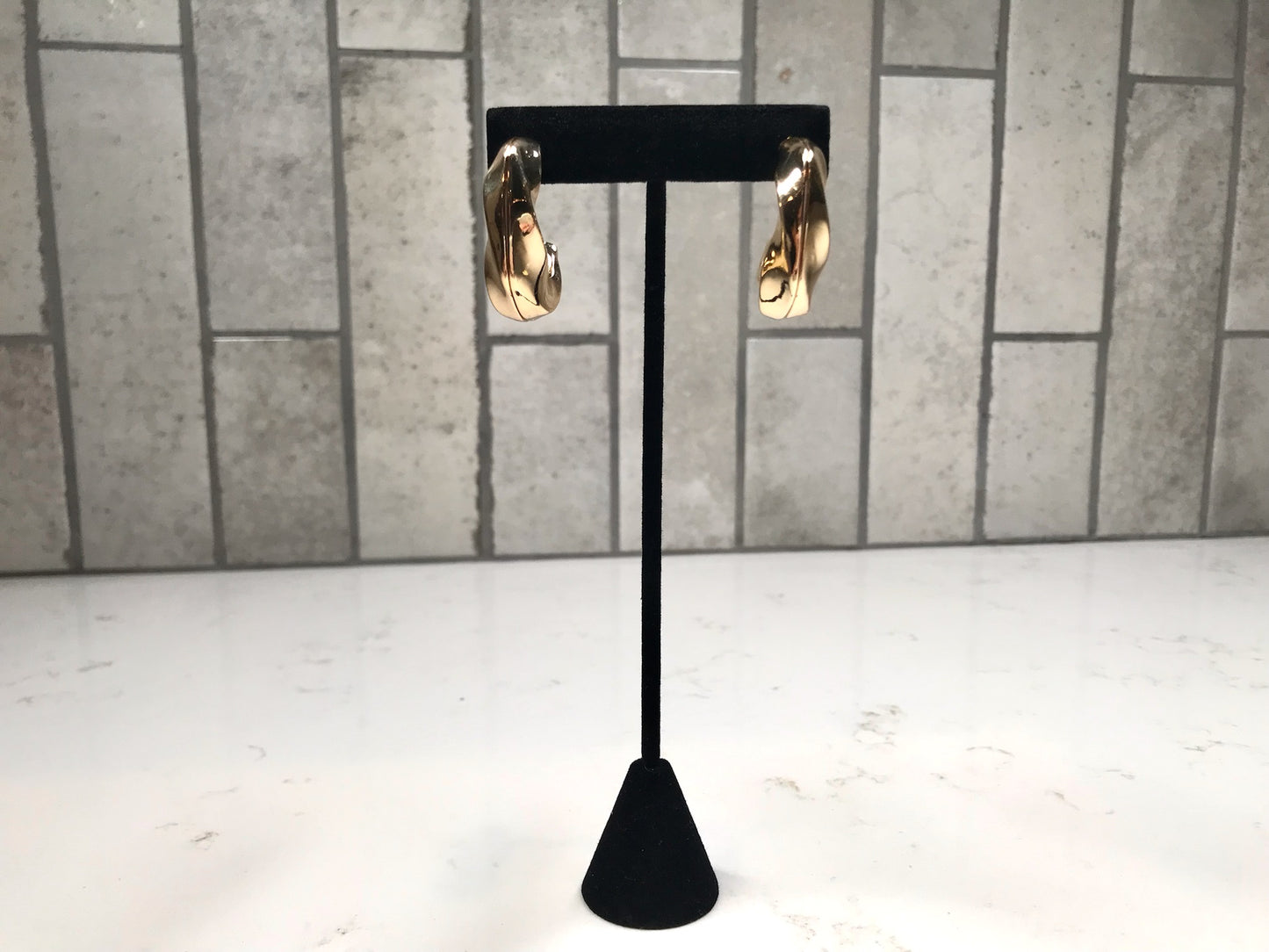 Gold chunky curved hoop earrings displayed on a black stand against a light gray tile background.