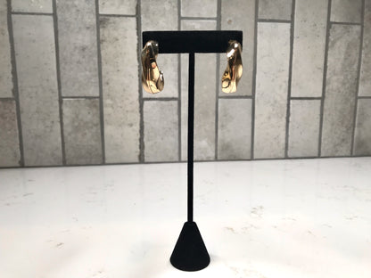 Gold chunky curved hoop earrings displayed on a black stand against a light gray tile background.