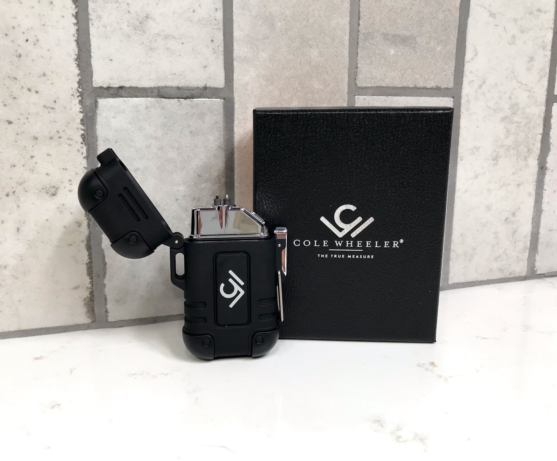 Black lighter with Cole Wheeler logo on a black box against a tiled wall background