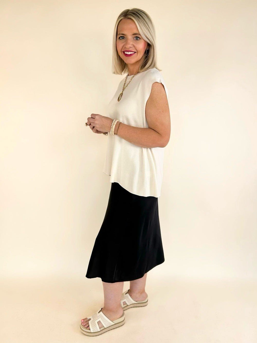 Women's ivory sleeveless flowy blouse with gold button shoulder accents, relaxed fit, and high-low hem styled with a black midi skirt.