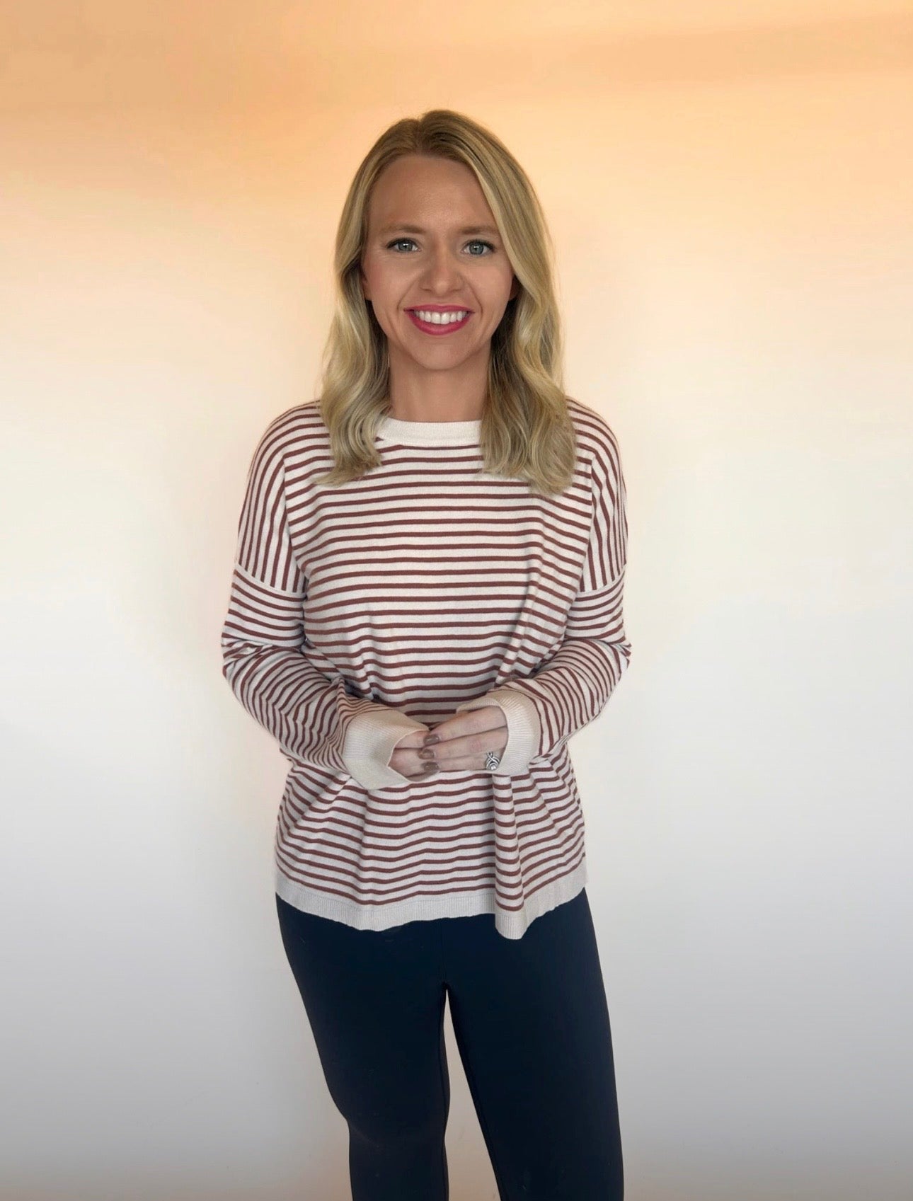 Woman wearing a cream and rust striped long-sleeve sweater with black pants, standing against a neutral beige backdrop. Casual fall outfit style.