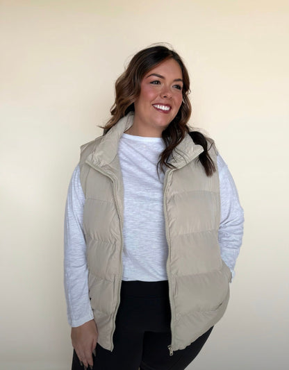 Woman wearing a light beige puffer vest over a white long-sleeve top and black leggings, smiling and looking to the side against a neutral background.