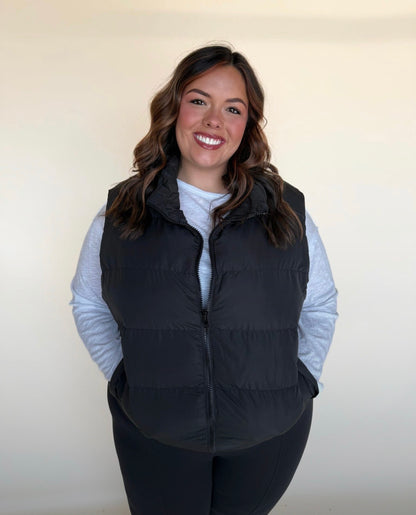 Woman wearing a black puffer vest over a white long-sleeve top and black leggings, hands in pockets, smiling brightly in front of a neutral backdrop.
