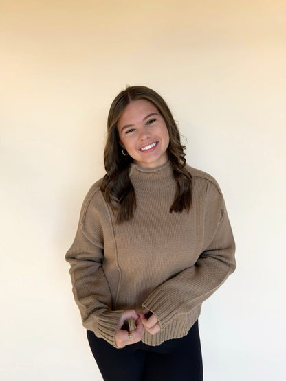 Woman wearing a taupe mock-neck knit sweater with ribbed cuffs and hem, styled with black leggings against a soft neutral background.