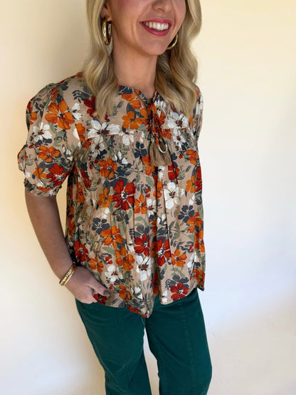 Close-up of a woman wearing a tan floral blouse with orange, white, and navy flowers, showing the tie neckline and gathered detail.