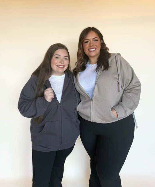 Two women wearing zip-up hoodies in charcoal and taupe, styled with white tops and black leggings, smiling in natural light.
