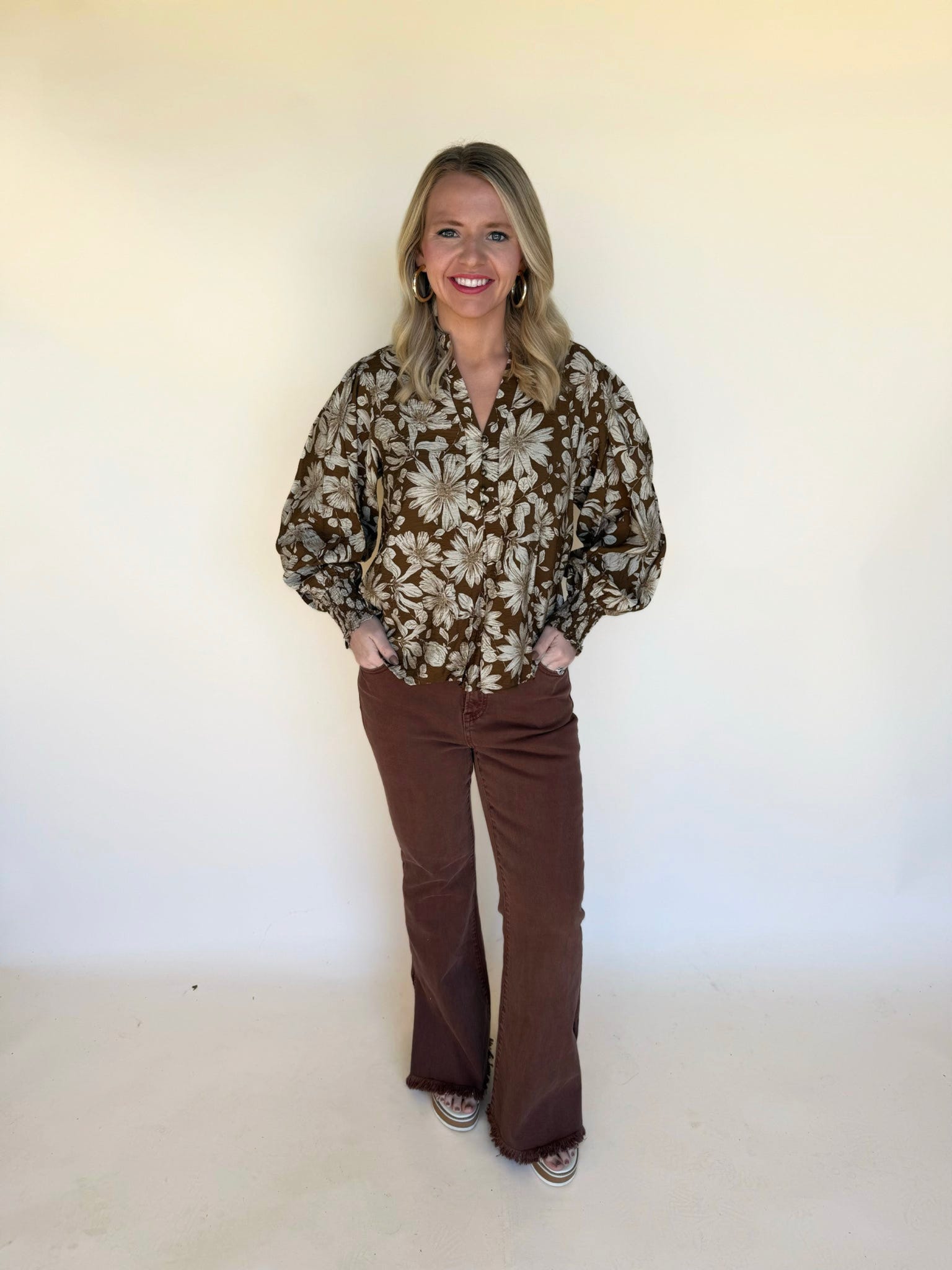 Woman wearing a brown floral blouse with balloon sleeves and brown flare pants, standing against a neutral background.