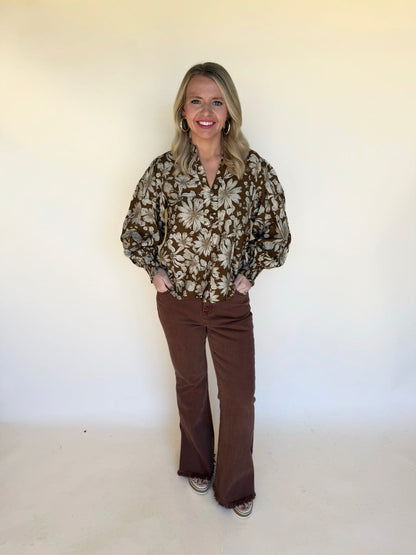 Woman wearing a brown floral blouse with balloon sleeves and brown flare pants, standing against a neutral background.