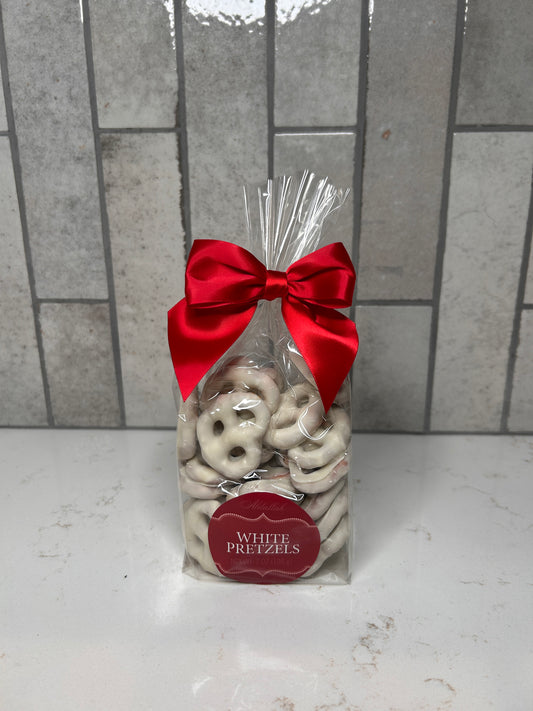 A clear gift bag filled with white chocolate–covered pretzels, tied with a red satin bow, sitting on a white countertop with a gray tile background.