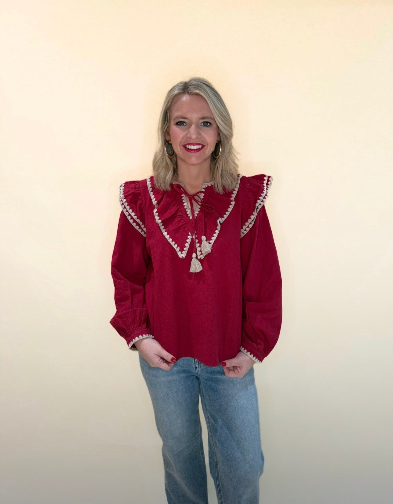 Front view of long sleeve red blouse with cream embroidered ruffle detail and tassel tie neckline, paired with light wash jeans.