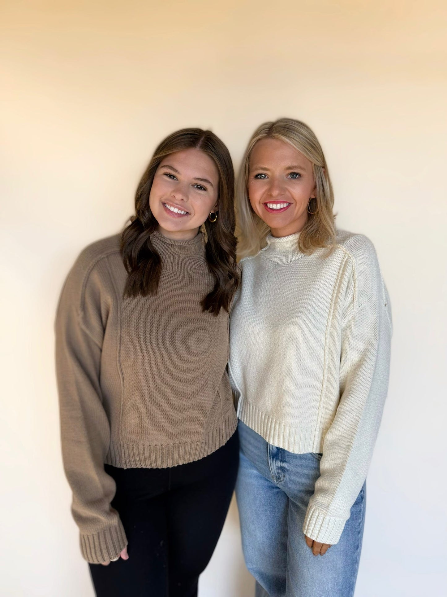Two women standing together wearing mock-neck knit sweaters; one in a taupe pullover with long sleeves and the other in an ivory knit sweater paired with blue jeans, smiling in front of a neutral background.