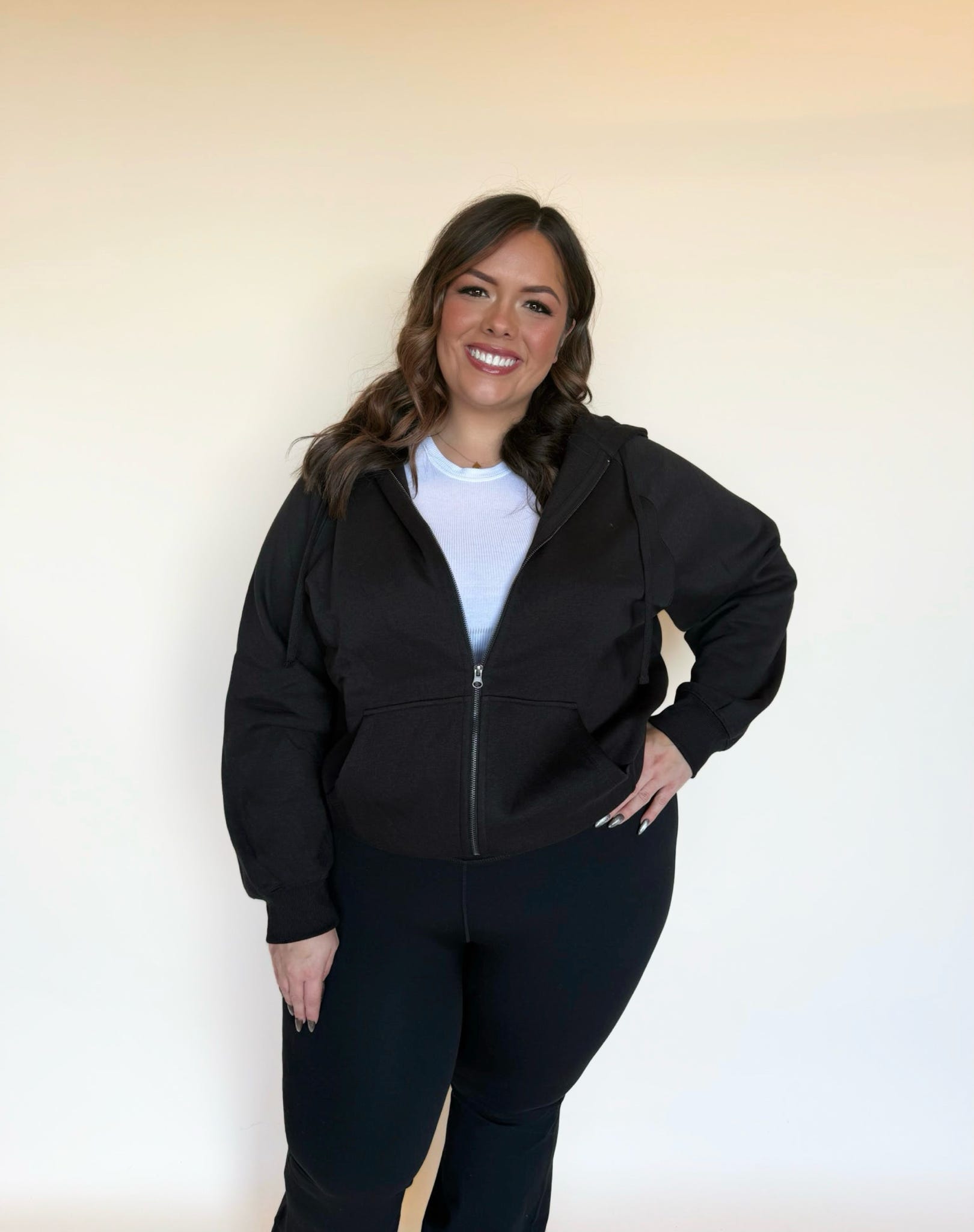 Woman wearing a black zip-up hoodie layered over a white top, smiling with one hand on her hip. 