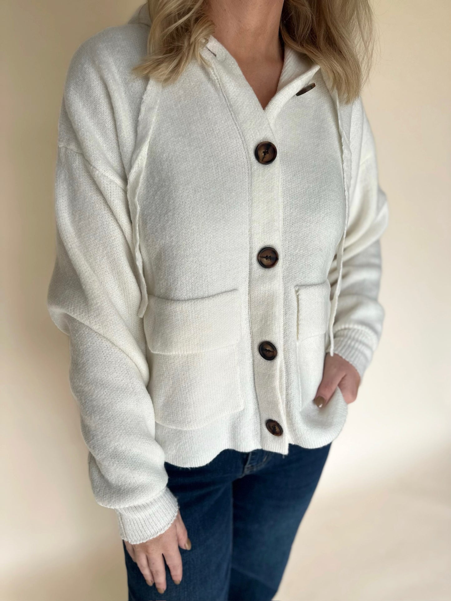 Close-up of a cream knit button-front hoodie cardigan featuring oversized tortoise buttons and two front patch pockets, paired with dark denim jeans.