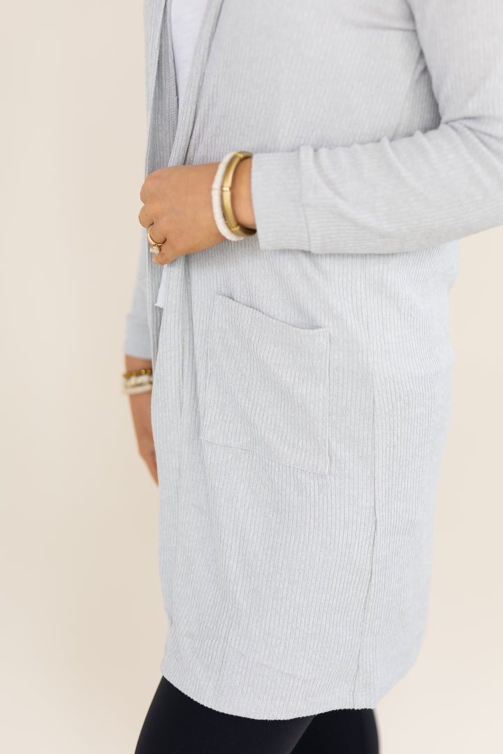 A close-up of a person wearing a light gray ribbed cardigan with a front patch pocket, paired with black leggings and gold-toned bracelets, against a plain beige background.