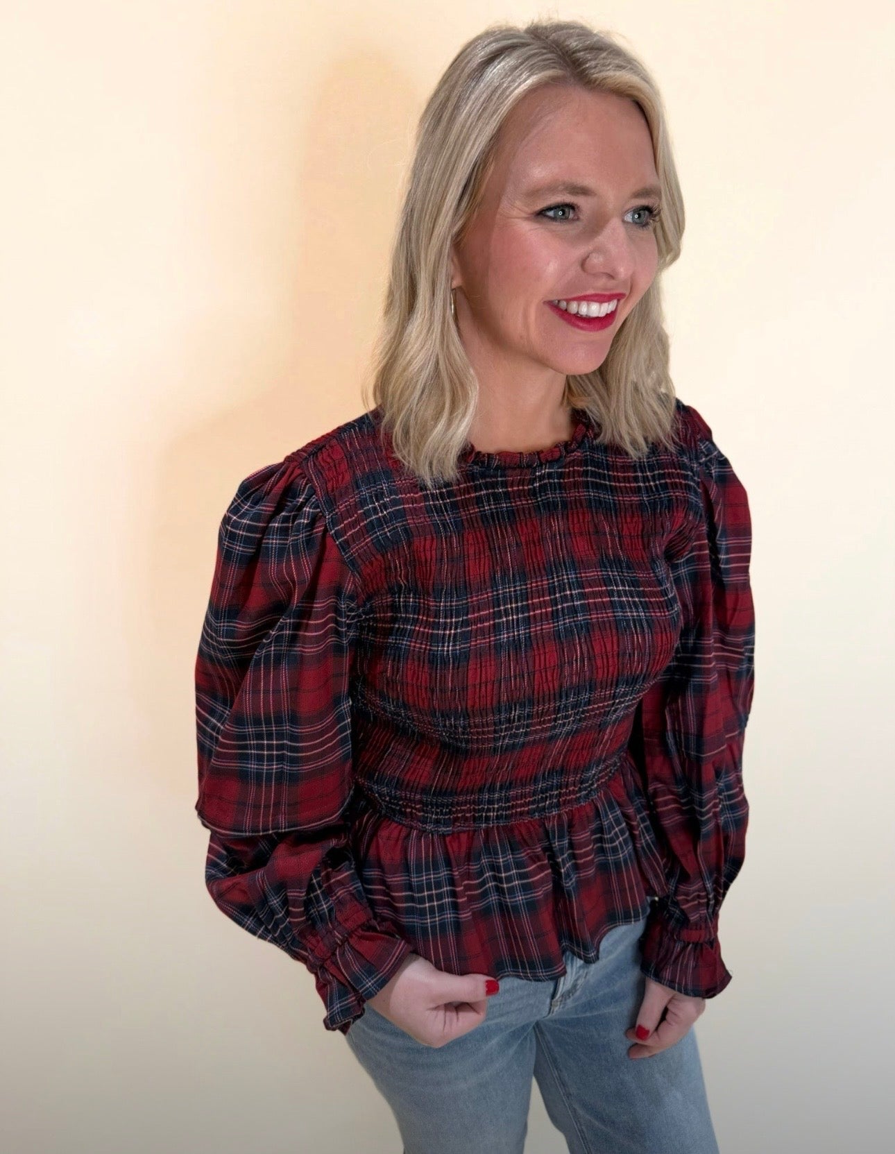 Red and navy plaid long sleeve top with smocked waist and puff sleeves shown from a front-side angle, styled with denim for a casual holiday look.