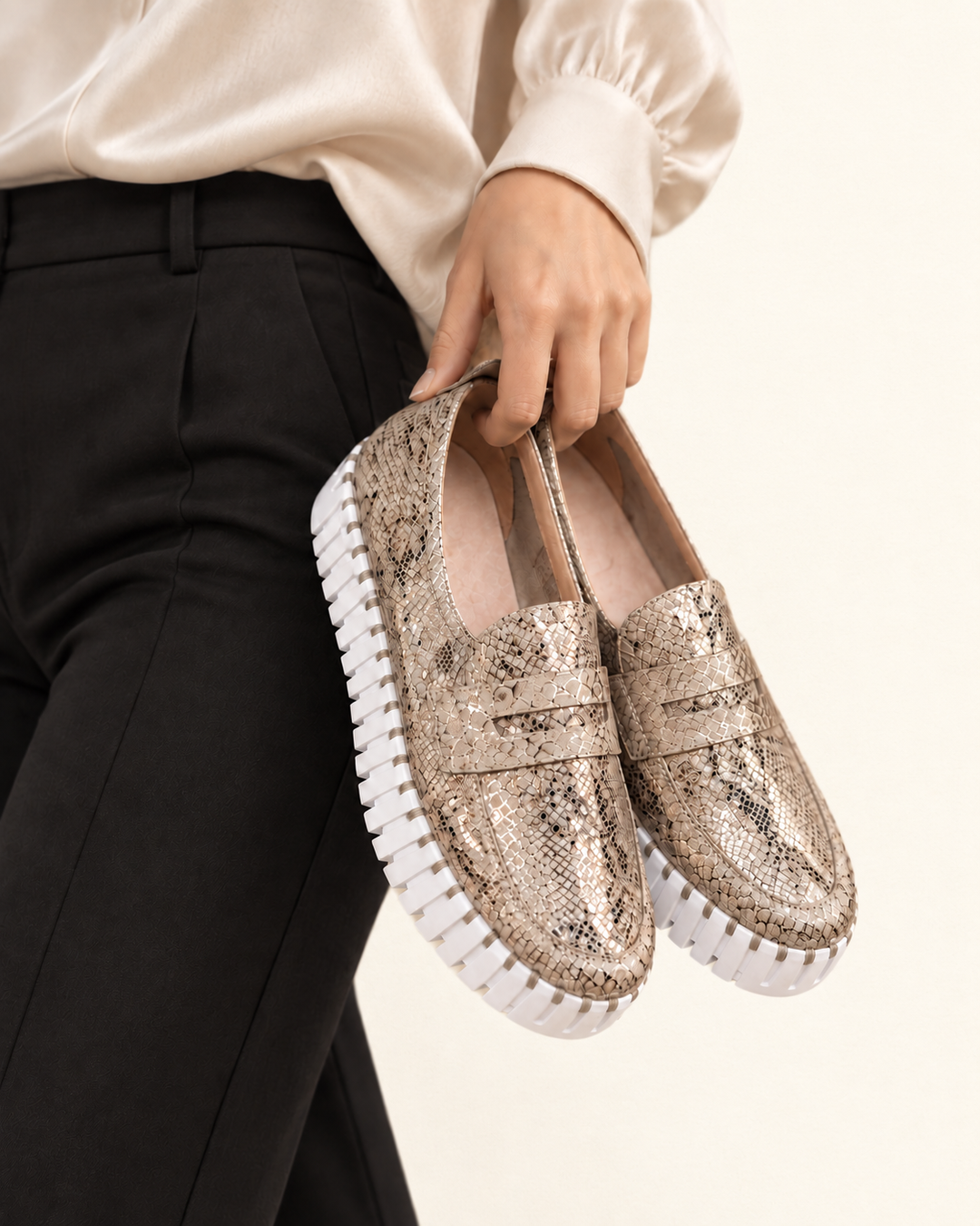 Woman wearing an ivory blouse and black trousers holding metallic champagne snakeskin platform loafers with white segmented soles against a soft ivory background.