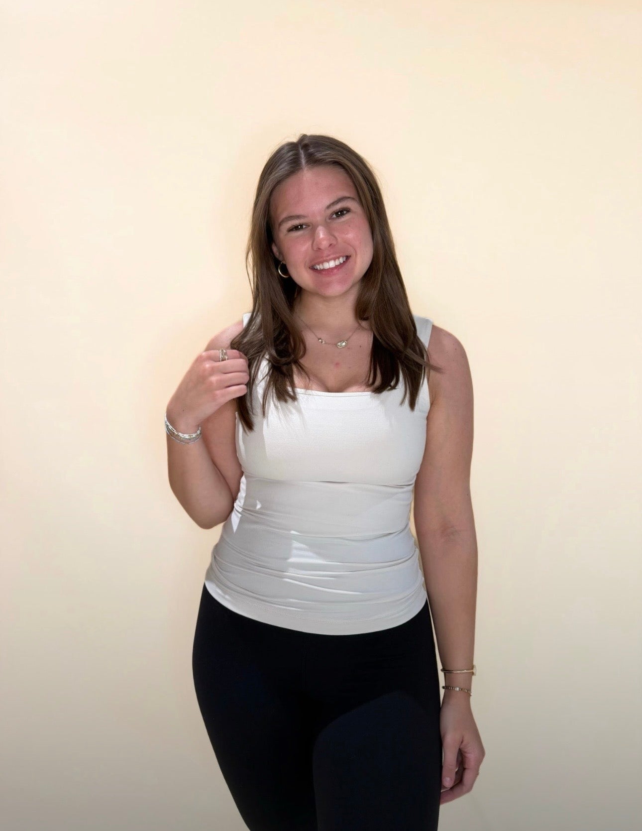 Young woman in a pearl white square-neck tank top and black leggings, smiling with long straight hair, against a soft beige backdrop.
