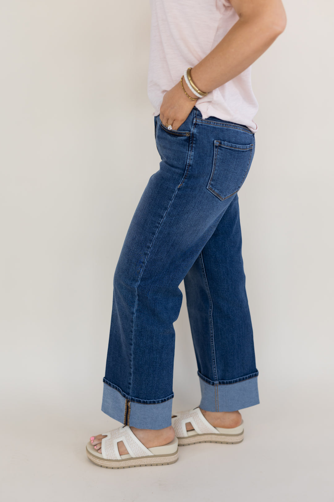 A person wearing blue wide-leg cuffed jeans, a light pink top, and white slide sandals, shown from the waist down against a plain light background.