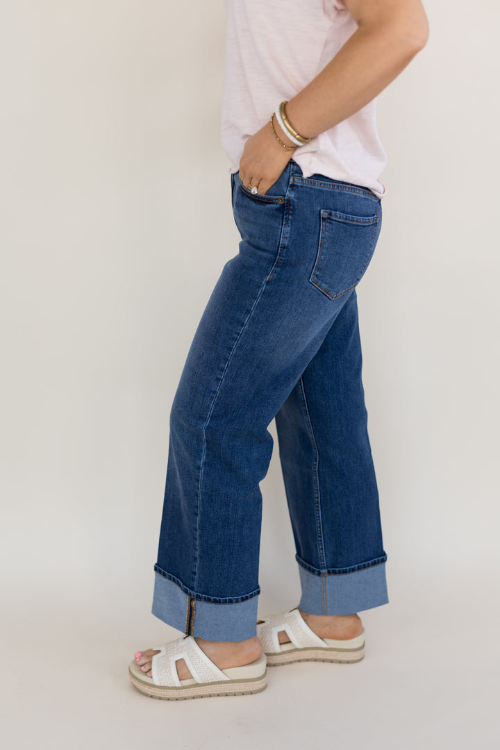 A person wearing blue wide-leg cuffed jeans, a light pink top, and white slide sandals, shown from the waist down against a plain light background.
