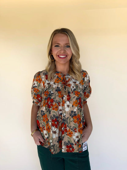 Woman wearing a taupe floral babydoll top with rust, orange, navy, and white flowers, paired with dark green pants. Smocked chest, puff sleeves, and tie-neck tassel detail. 