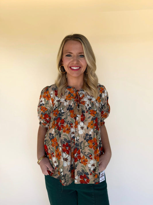 Woman wearing a taupe floral babydoll top with rust, orange, navy, and white flowers, paired with dark green pants. Smocked chest, puff sleeves, and tie-neck tassel detail. 