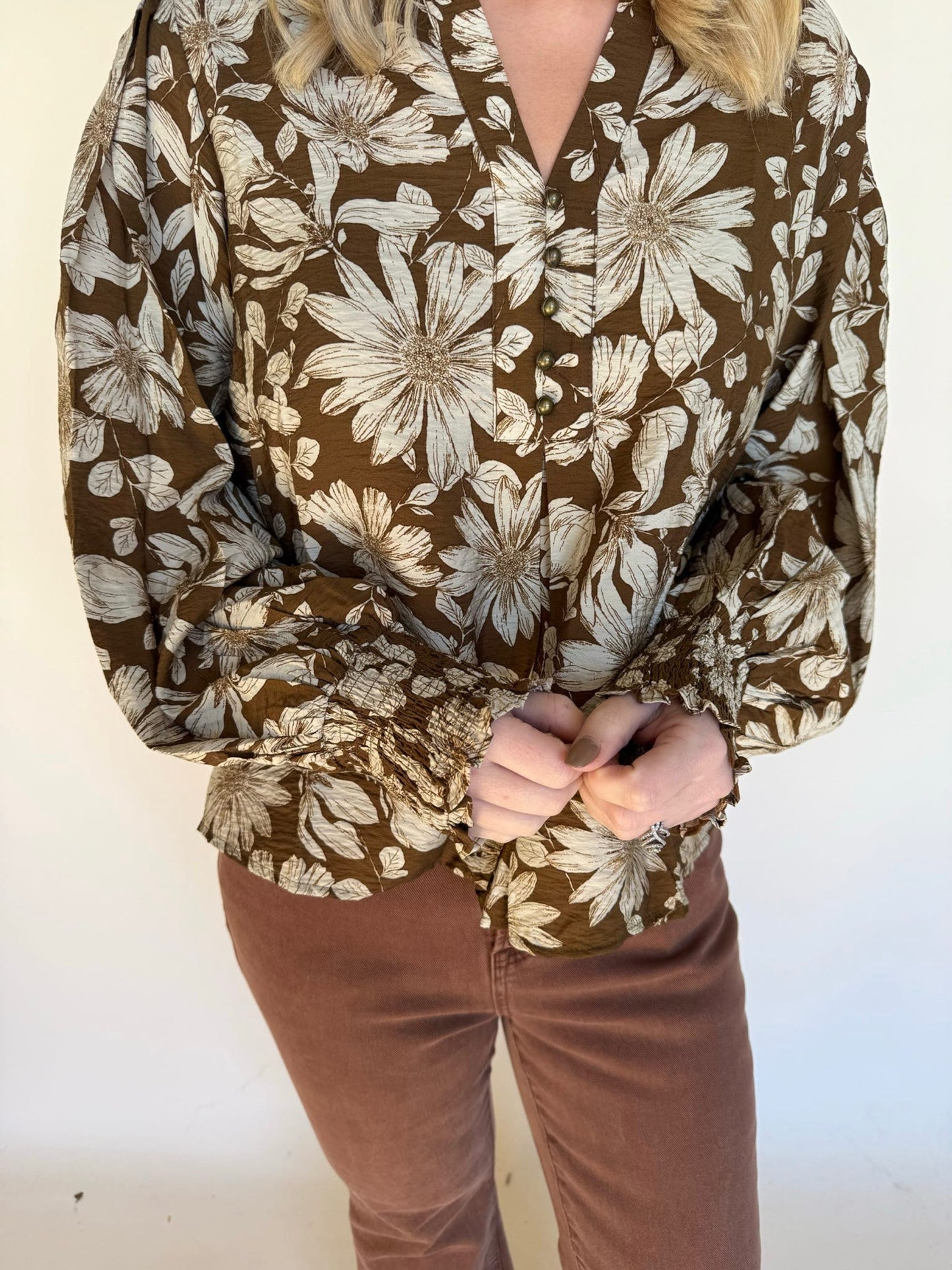 Detail shot of a brown floral blouse showing the button front, smocked sleeves, and textured fabric paired with brown pants.
