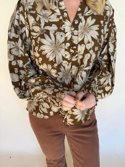 Detail shot of a brown floral blouse showing the button front, smocked sleeves, and textured fabric paired with brown pants.
