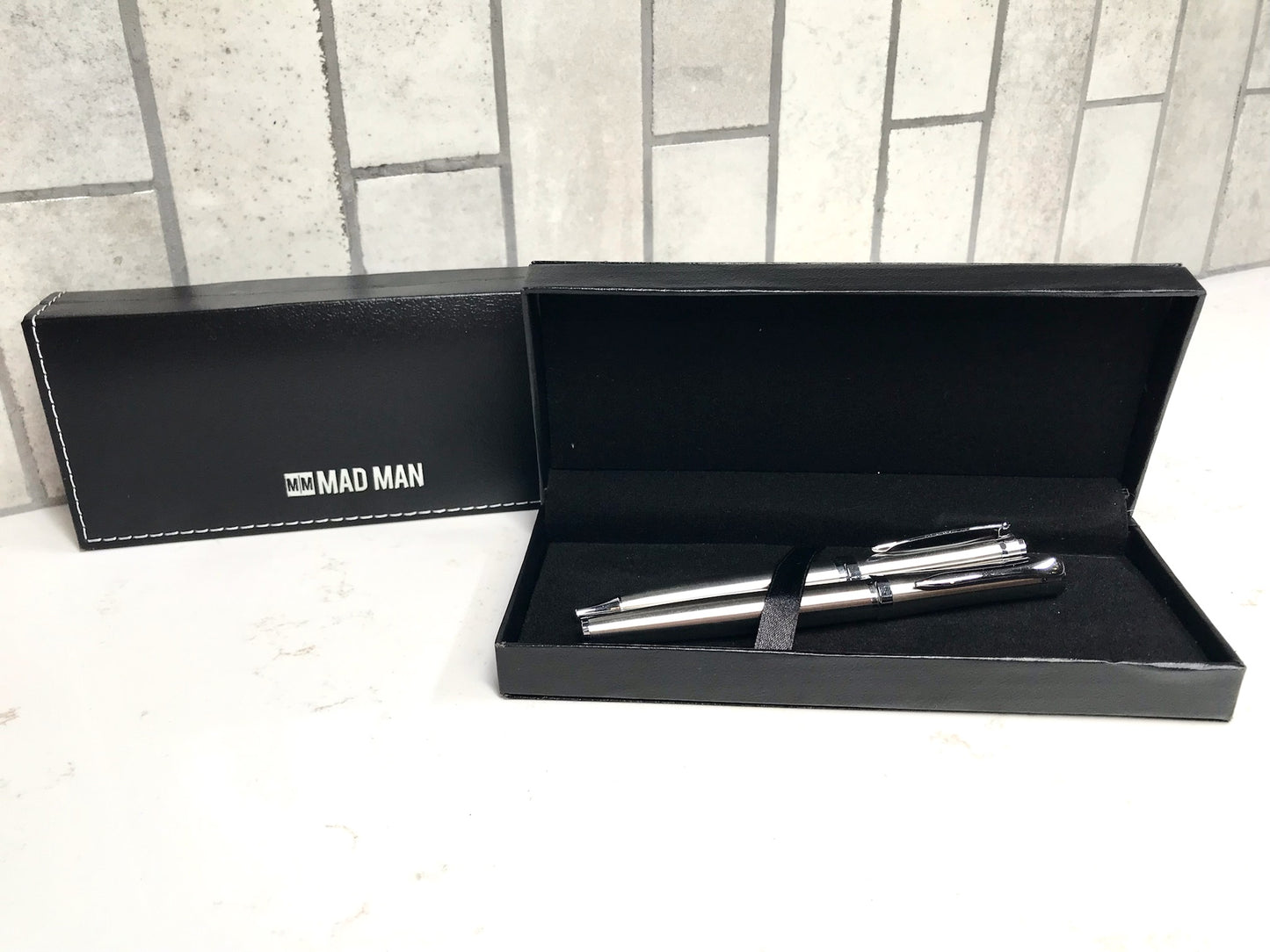 Two silver pens in a black box with 'MAD MAN' branding on a tiled floor.