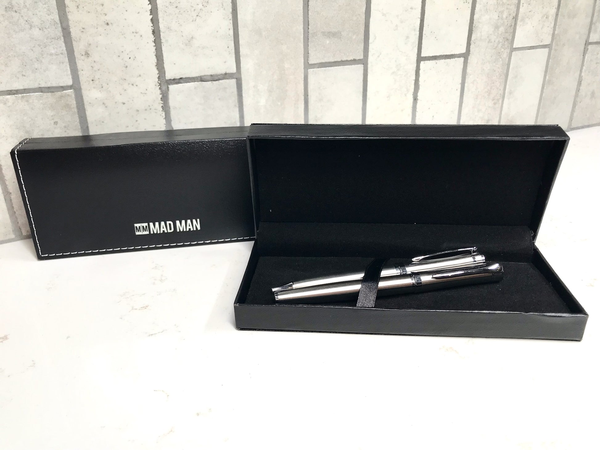 Two silver pens in a black box with 'MAD MAN' branding on a tiled floor.