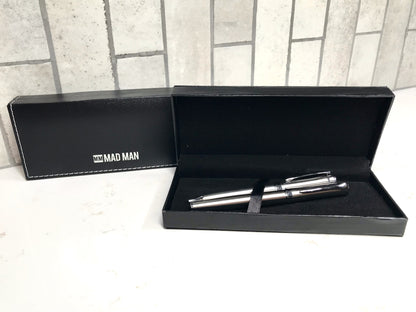 Two silver pens in a black box with 'MAD MAN' branding on a tiled floor.
