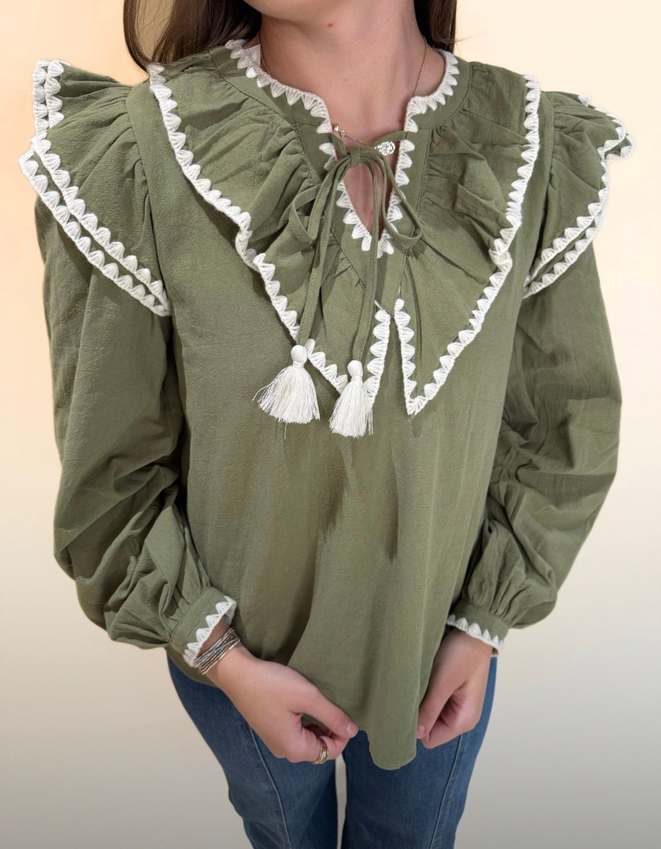 Close up of olive green blouse featuring white scalloped trim, ruffled yoke, and tassel neckline, styled with denim.