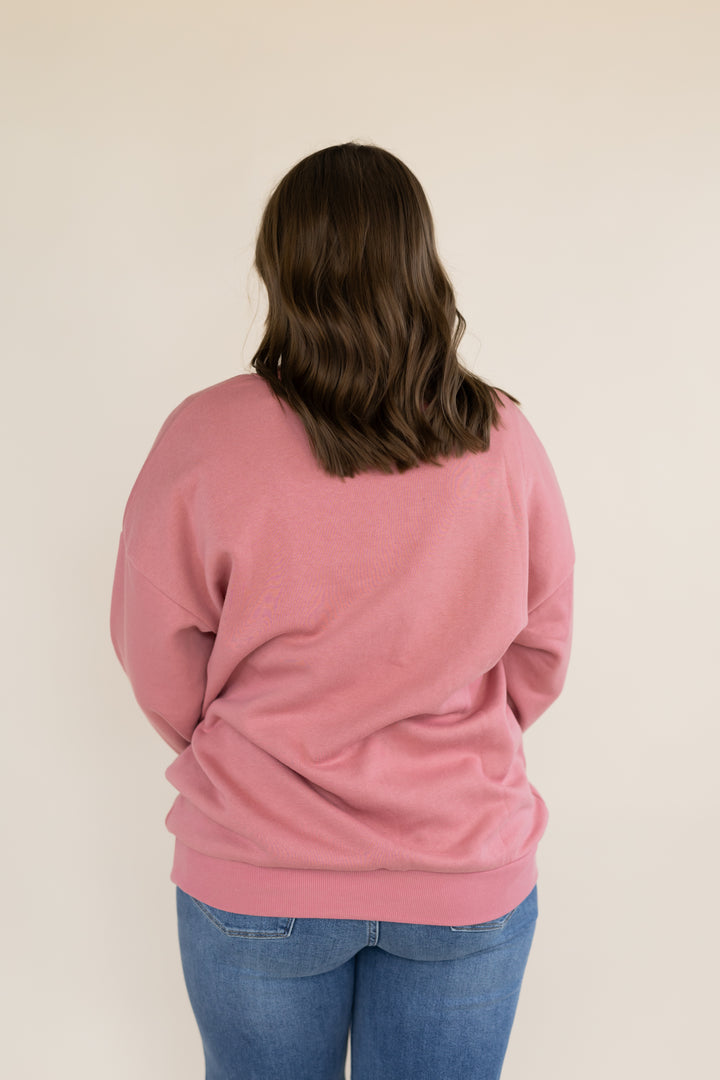 A person viewed from behind wearing a dusty pink crewneck sweatshirt and blue jeans, standing against a plain beige background.