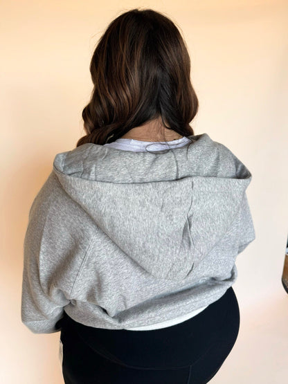 Back view of a woman wearing a light gray zip-up hoodie, showing the hood and relaxed fit.