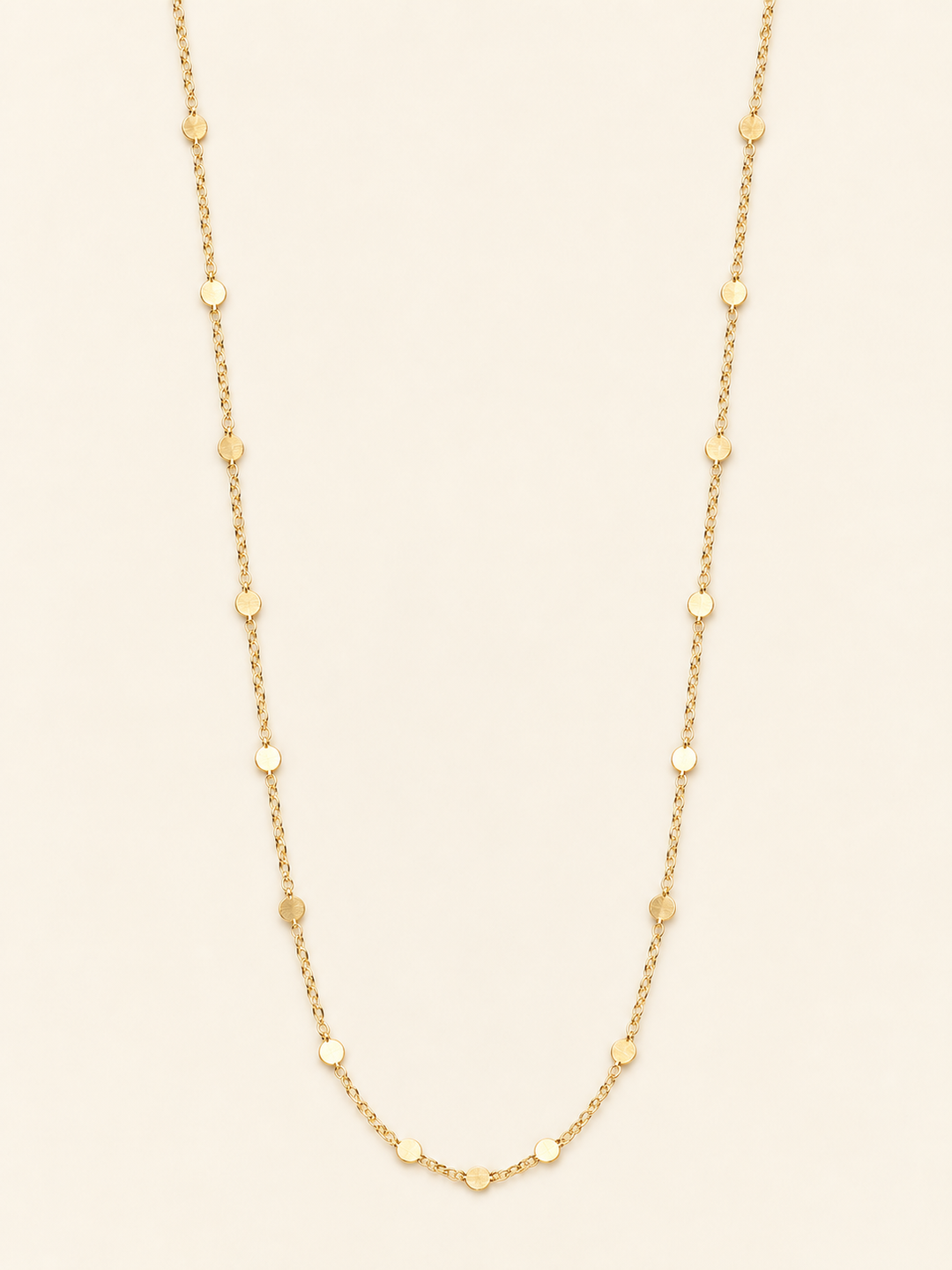 Long gold flat disc station chain necklace arranged on a cream studio background, featuring delicate chain links and evenly spaced polished round accents.