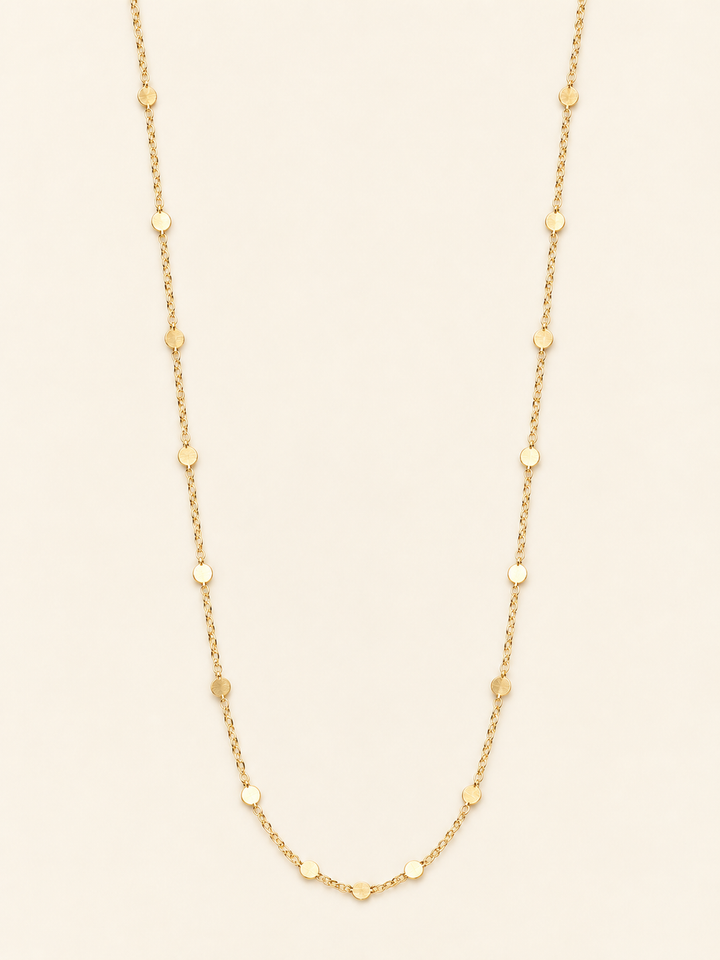 Long gold flat disc station chain necklace arranged on a cream studio background, featuring delicate chain links and evenly spaced polished round accents.